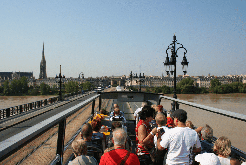 Photographie officielle : passagers à l’étage supérieur d’un bus touristique traversant le pont de pierre à Bordeaux.